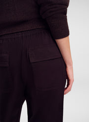 Close-up of a person wearing dark brown pants with a large front patch pocket and an elastic waistband, paired with a matching dark ribbed sweater, against a light gray studio background.