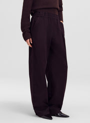 A model is shown from the waist down wearing dark brown wide-leg trousers, a matching dark brown knit sweater, and black pointed shoes against a light gray studio background.
