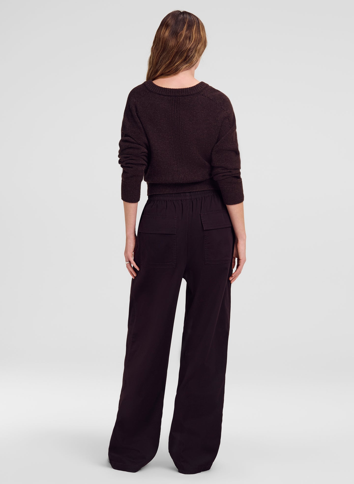 A woman viewed from the back wearing a brown knit sweater and matching wide-leg trousers against a light gray studio background.