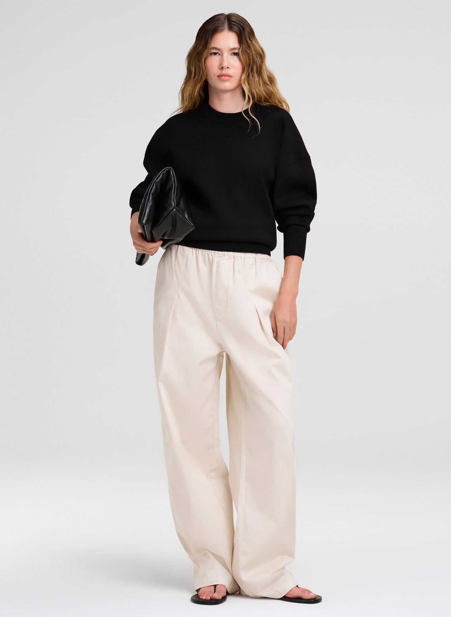 Woman wearing a black sweater and white pants holding a black clutch against a white background