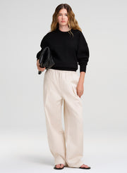 Woman wearing a black sweater and white pants holding a black clutch against a white background