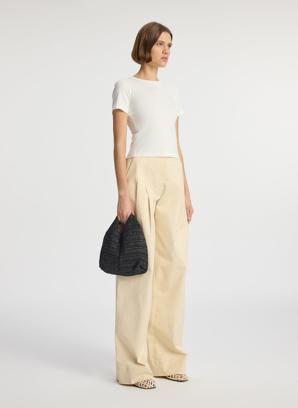 side view of woman in white tee and beige wide leg pants carrying small raffia woven handbag in black