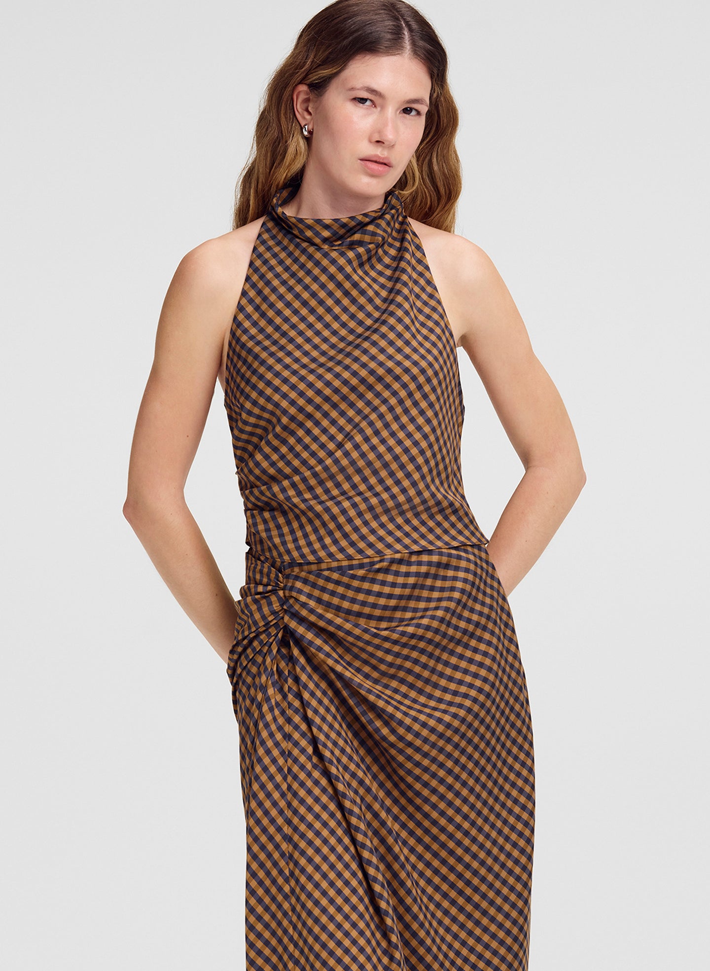 A woman modeling a sleeveless top with a brown and black checkered pattern, featuring a gathered ruched waist and a draped wrap-style skirt, set against a solid light gray background.