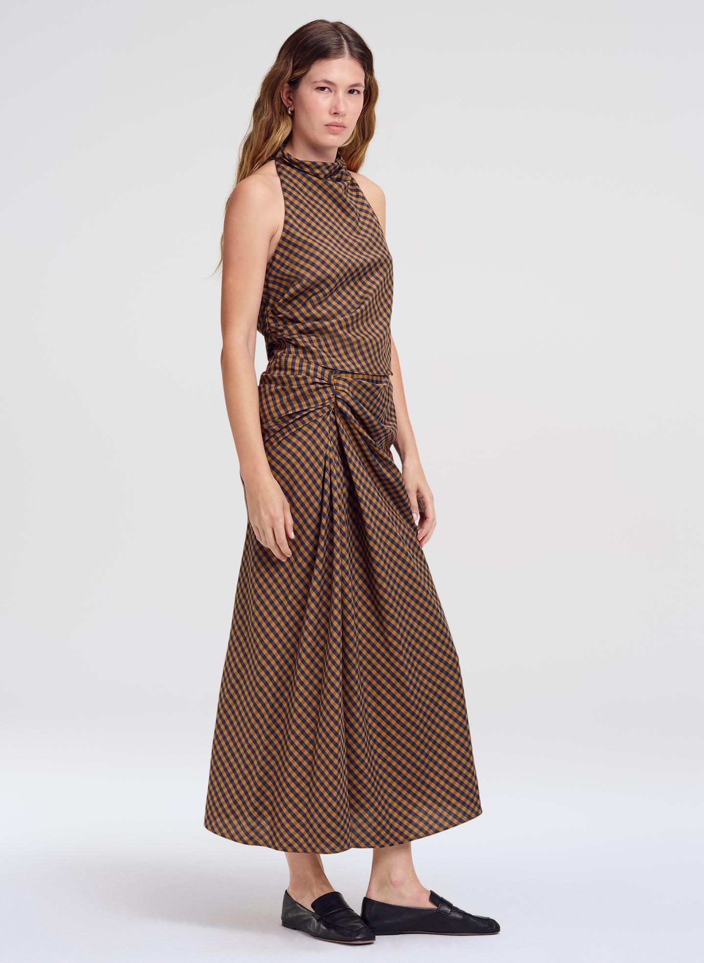 A woman modeling a sleeveless top and matching midi skirt with a brown and beige geometric pattern, styled with black slip-on shoes against a light gray studio background.