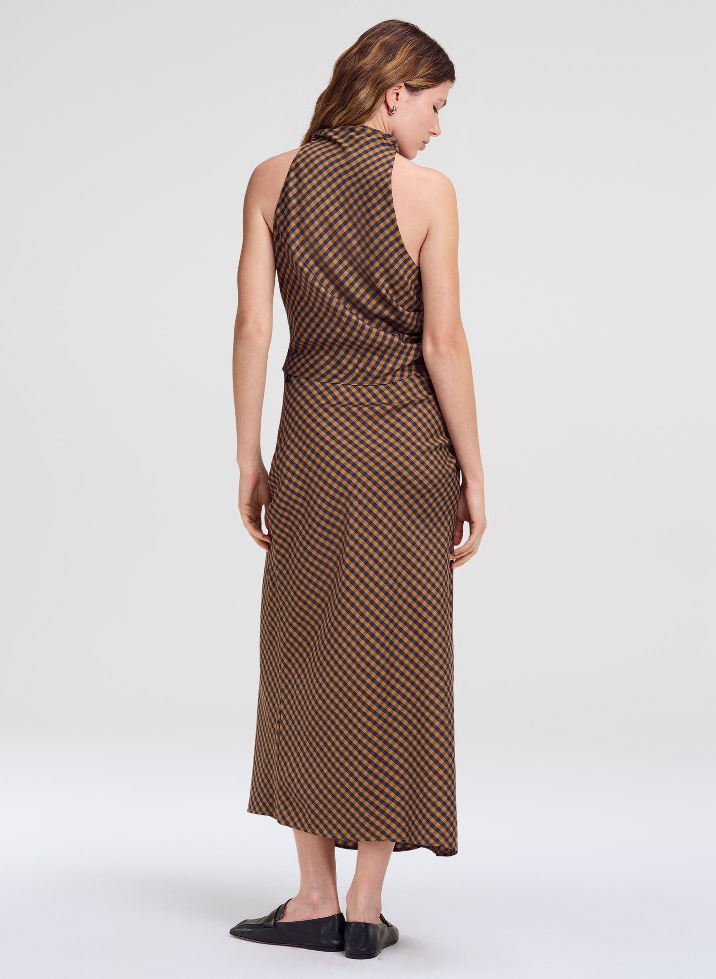 A woman shown from the back wearing a sleeveless brown outfit with a small repeating geometric pattern, photographed against a plain light gray studio background.