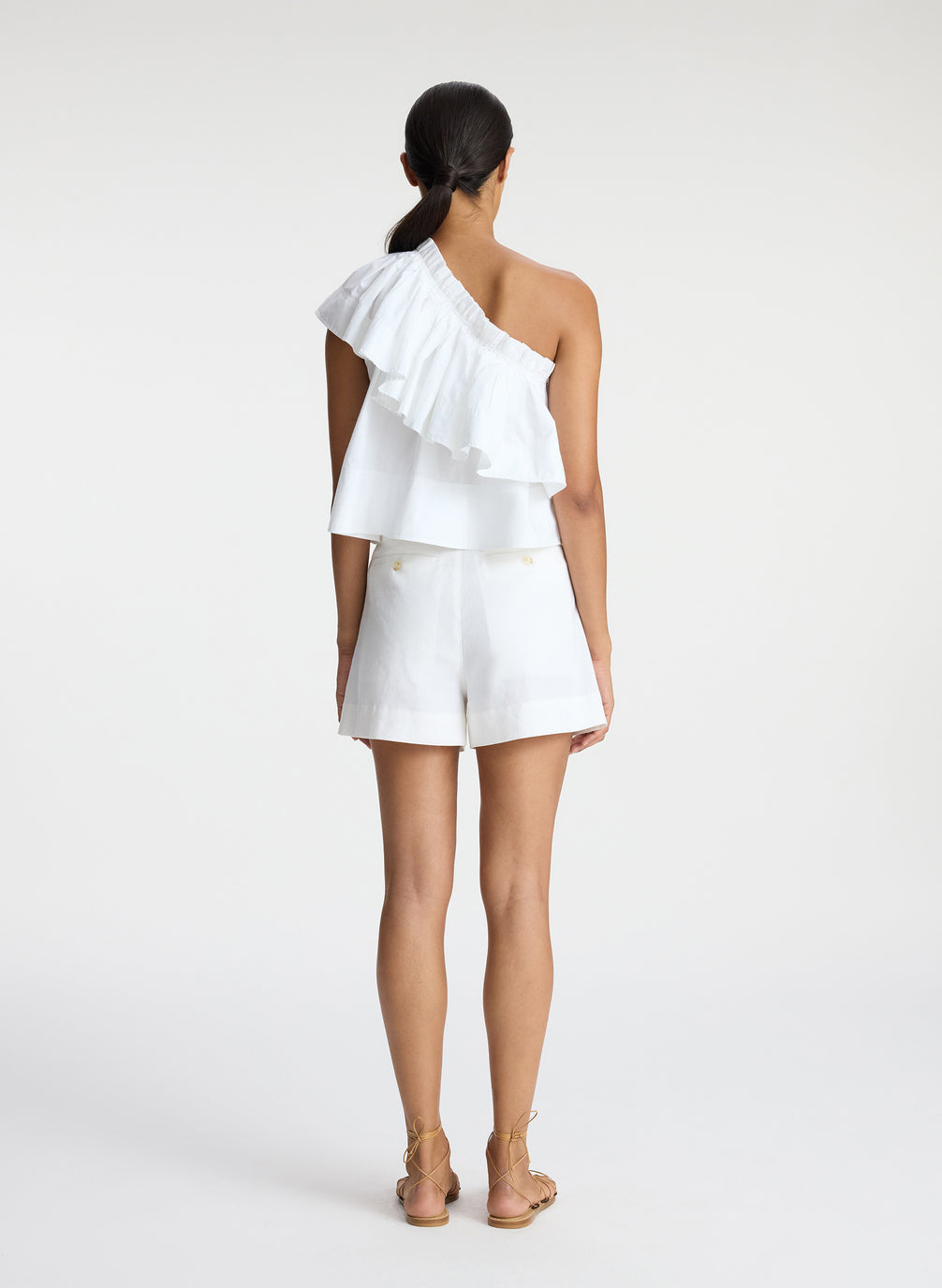 back view of woman wearing white one shoulder ruffle top and white shorts