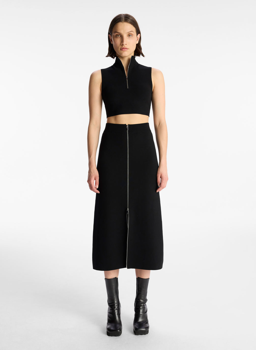 front view of woman wearing black half zip compact knit sleevless cropped top with matching black skirt