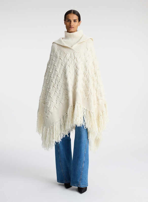 Front view of woman wearing cream cable knit poncho with fringe