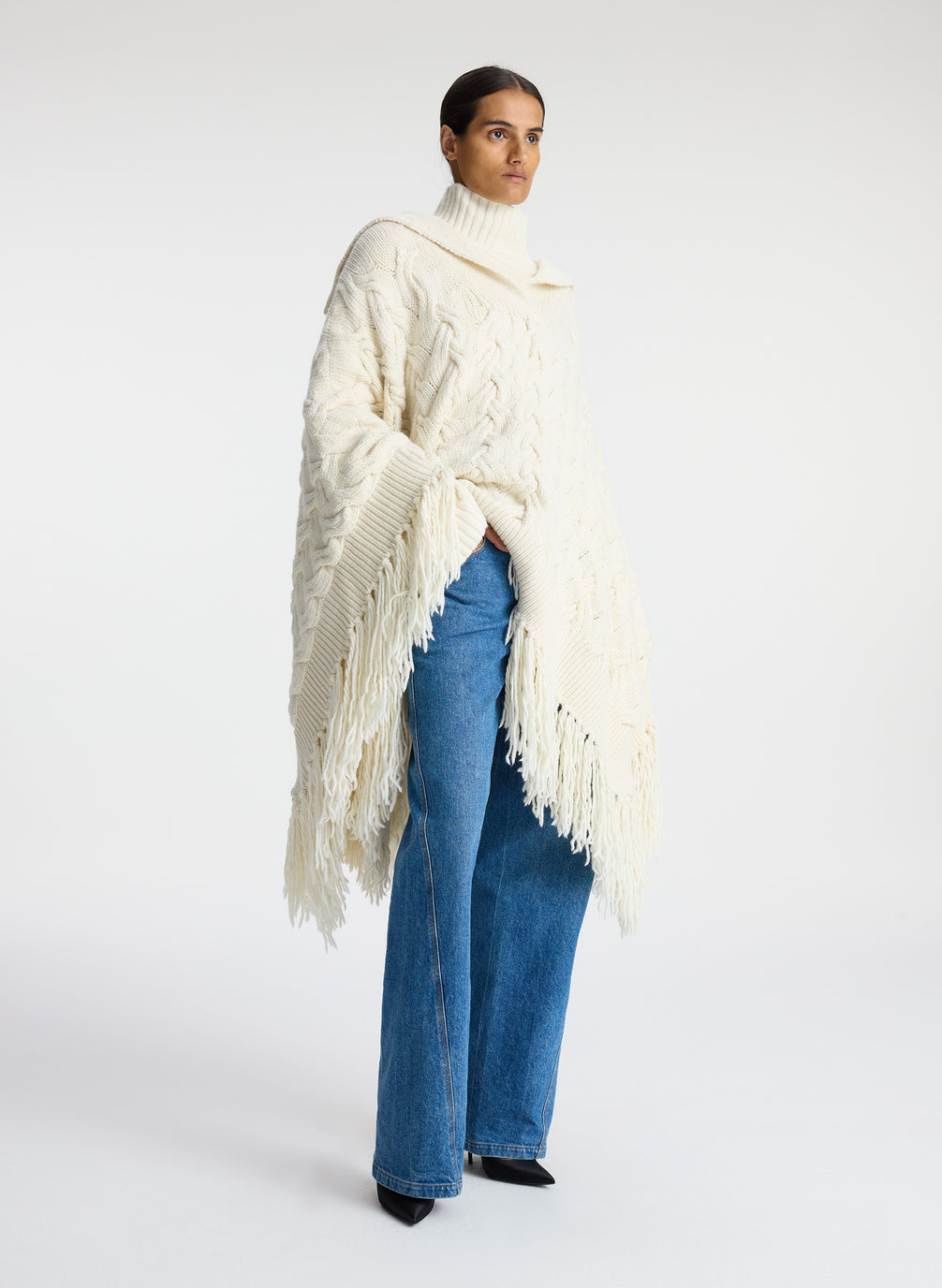 Side view of woman wearing cream cable knit poncho with fringe