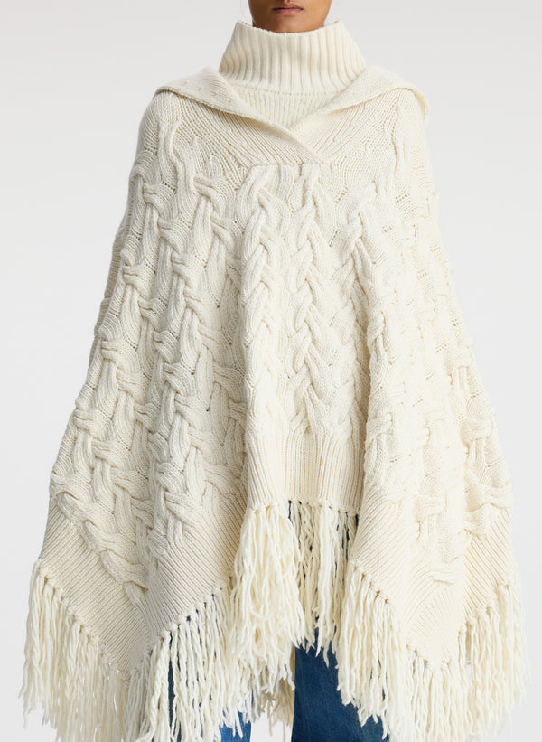 Front view of woman wearing cream cable knit poncho with fringe