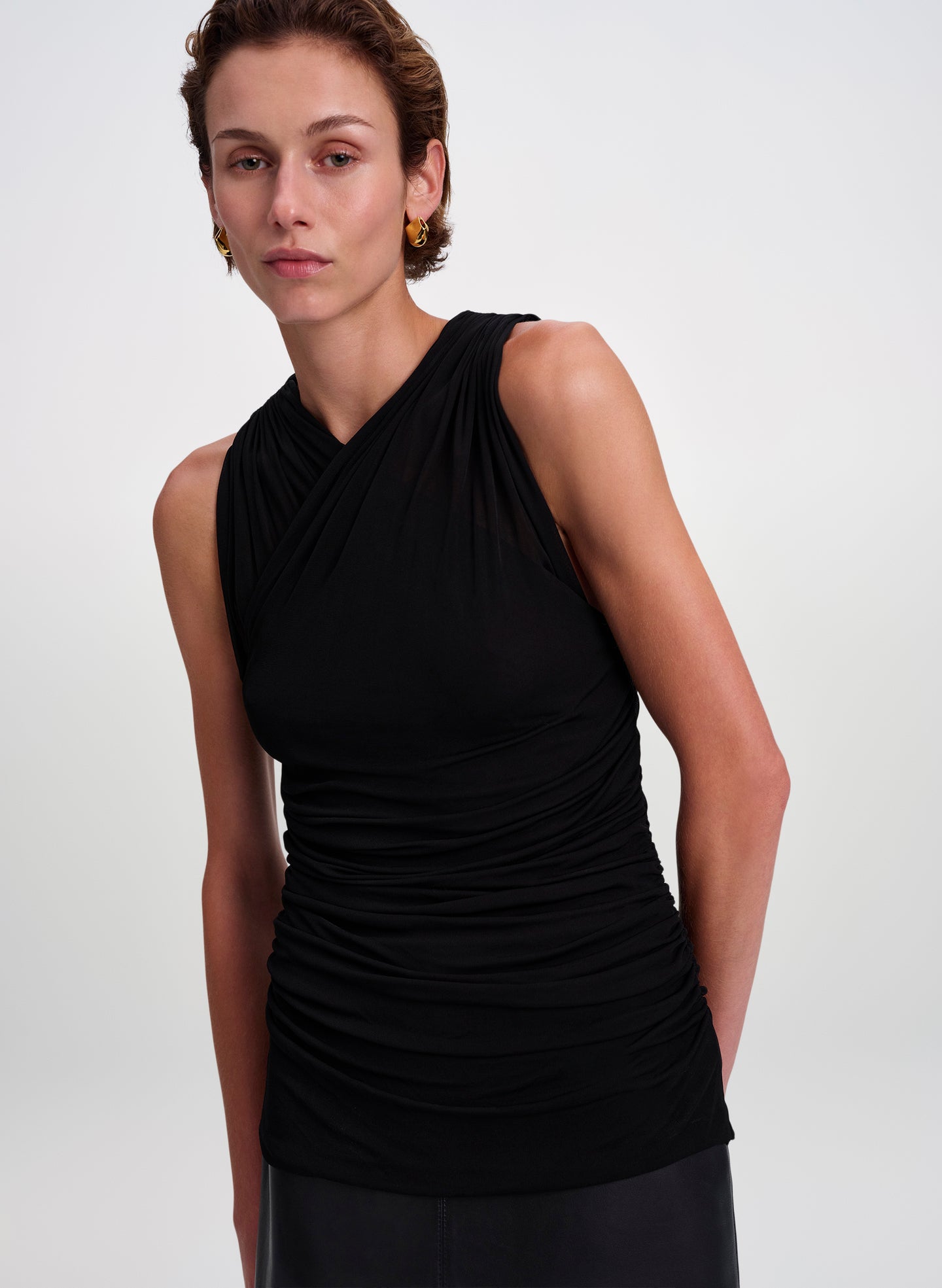 A woman modeling a black sleeveless ruched top with a crossover V-neckline, photographed against a clean light gray studio background.