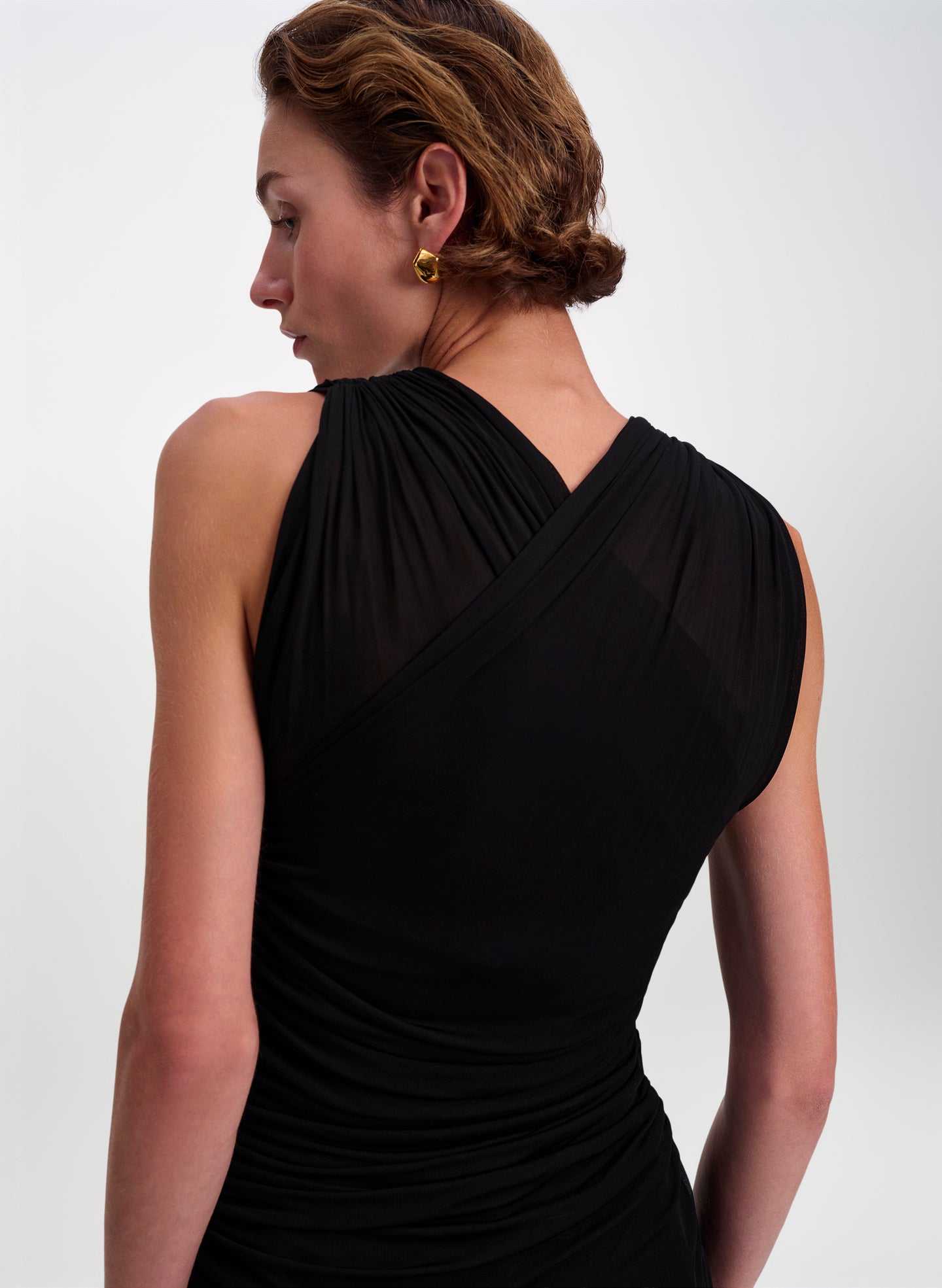 A woman shown from the back wearing a black draped sleeveless dress with sheer mesh paneling, photographed against a clean light gray studio background.