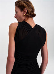 A woman shown from the back wearing a black draped sleeveless dress with sheer mesh paneling, photographed against a clean light gray studio background.