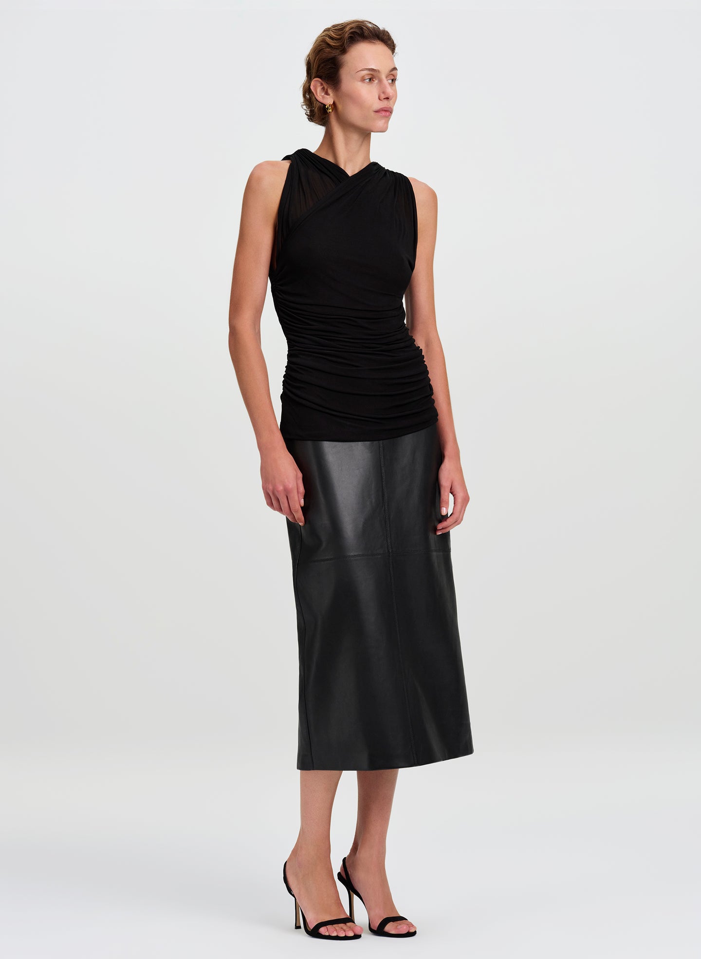 A model wearing a sleeveless black top with a ruched, draped bodice and a matching black faux-leather midi skirt, paired with black high-heeled sandals, against a light gray studio background.