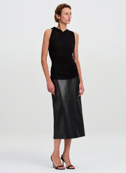 A model wearing a sleeveless black top with a ruched, draped bodice and a matching black faux-leather midi skirt, paired with black high-heeled sandals, against a light gray studio background.
