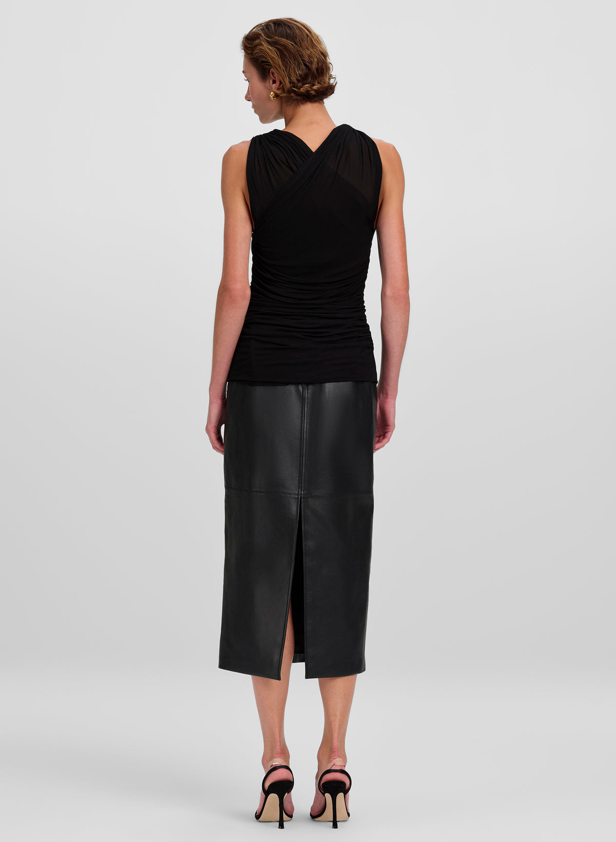 A woman shown from the back wearing a black sleeveless ruched top and a black leather midi skirt with a slit, paired with black high-heeled sandals, against a light gray studio background.