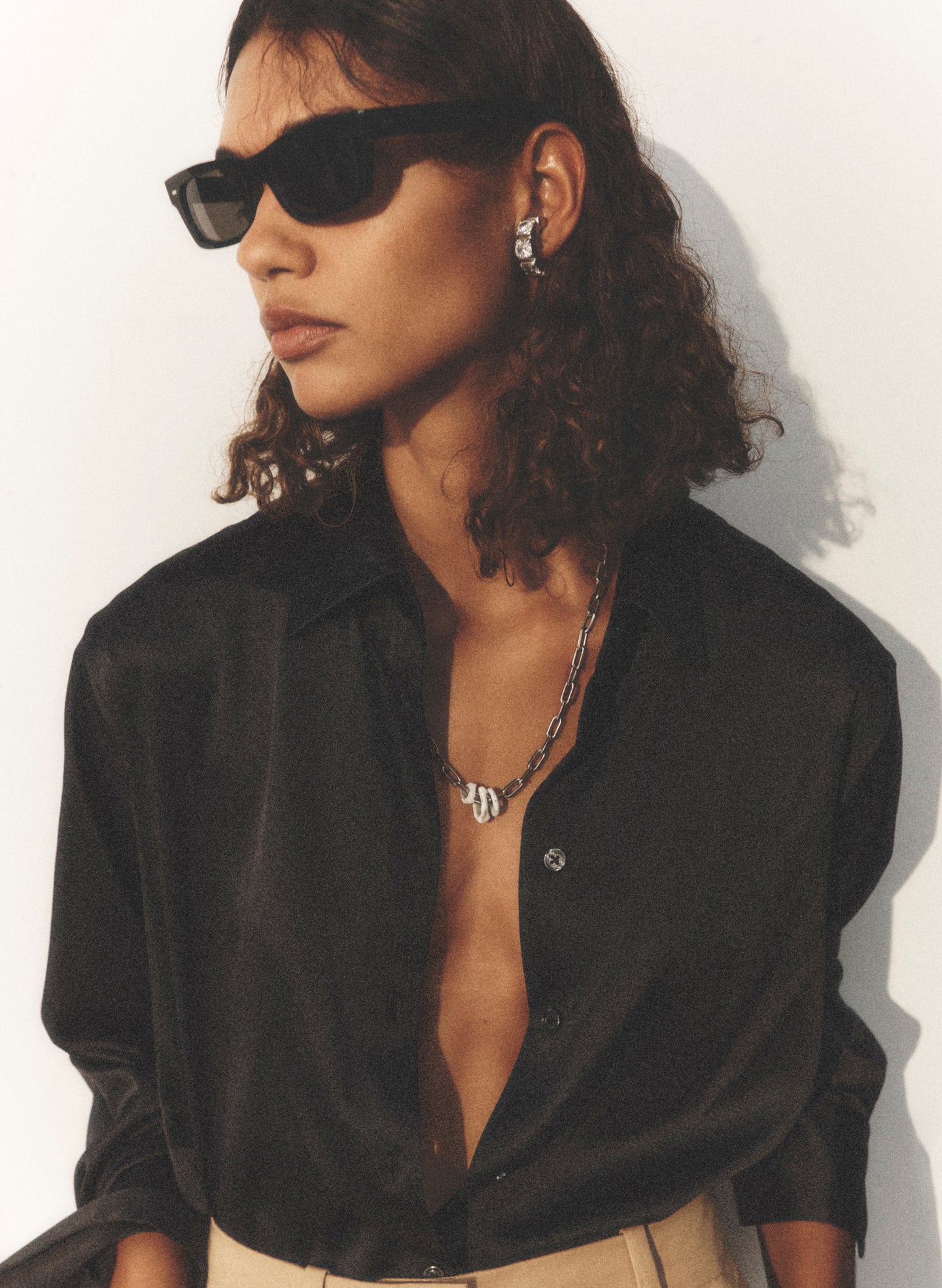 A woman wearing black sunglasses, a black silk button-up shirt, and beige trousers, photographed against a plain white wall with strong sunlight and shadows.