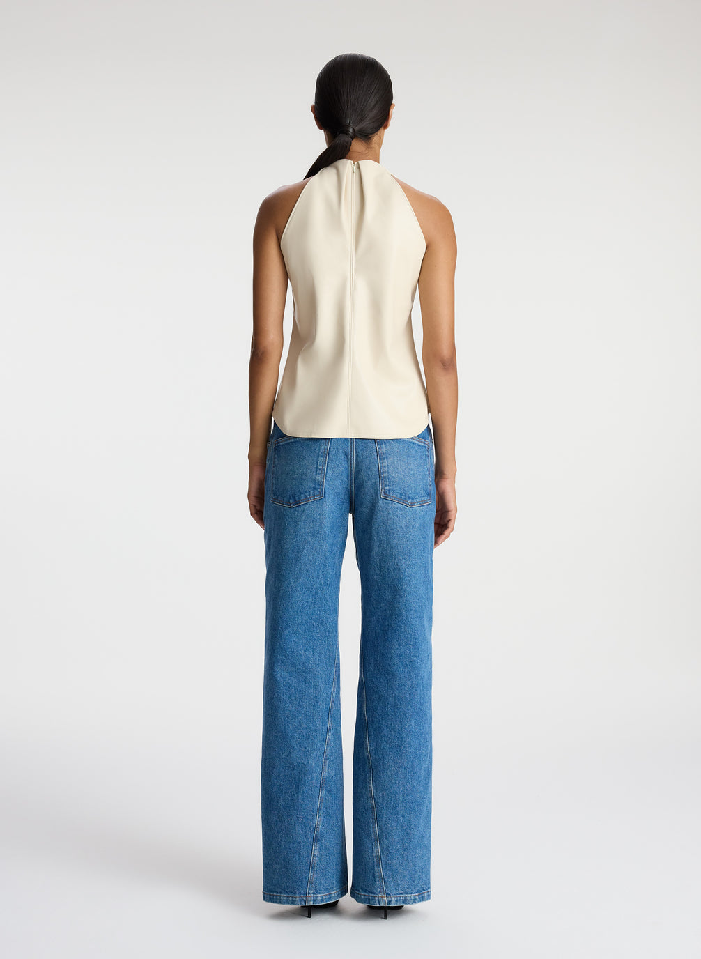 back view of woman wearing cream vegan leather halter top and medium blue wash denim jeans