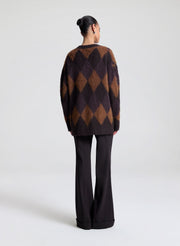 Person wearing a brown and black diamond pattern sweater with black pants on a white background