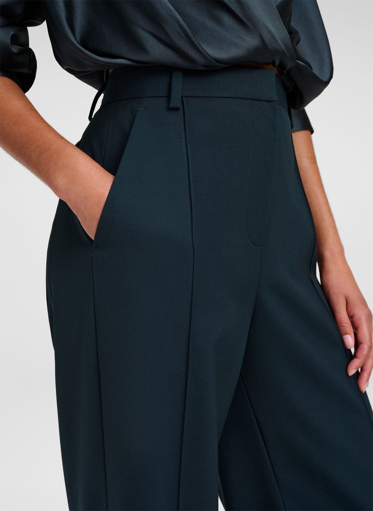 Close-up of a model wearing dark green wide-leg trousers with a high waist, belt, and side pockets, paired with a dark satin blouse against a light gray background.