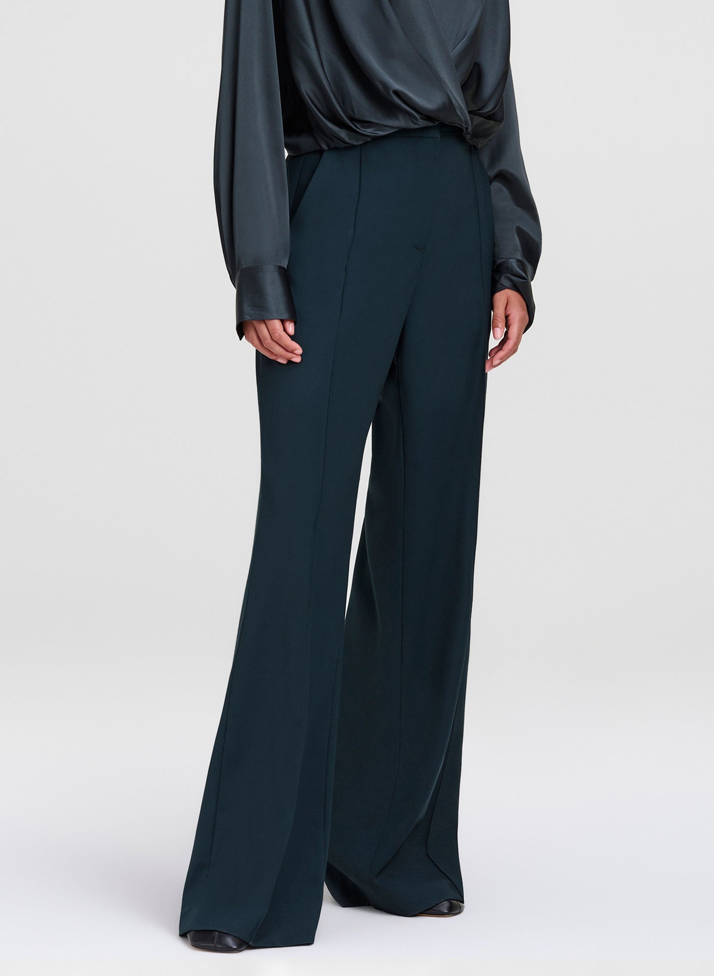 A model wearing dark teal wide-leg trousers and a matching dark satin blouse, shown from the chest down against a light gray studio background.