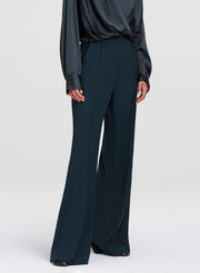 A model wearing dark teal wide-leg trousers and a matching dark satin blouse, shown from the chest down against a light gray studio background.