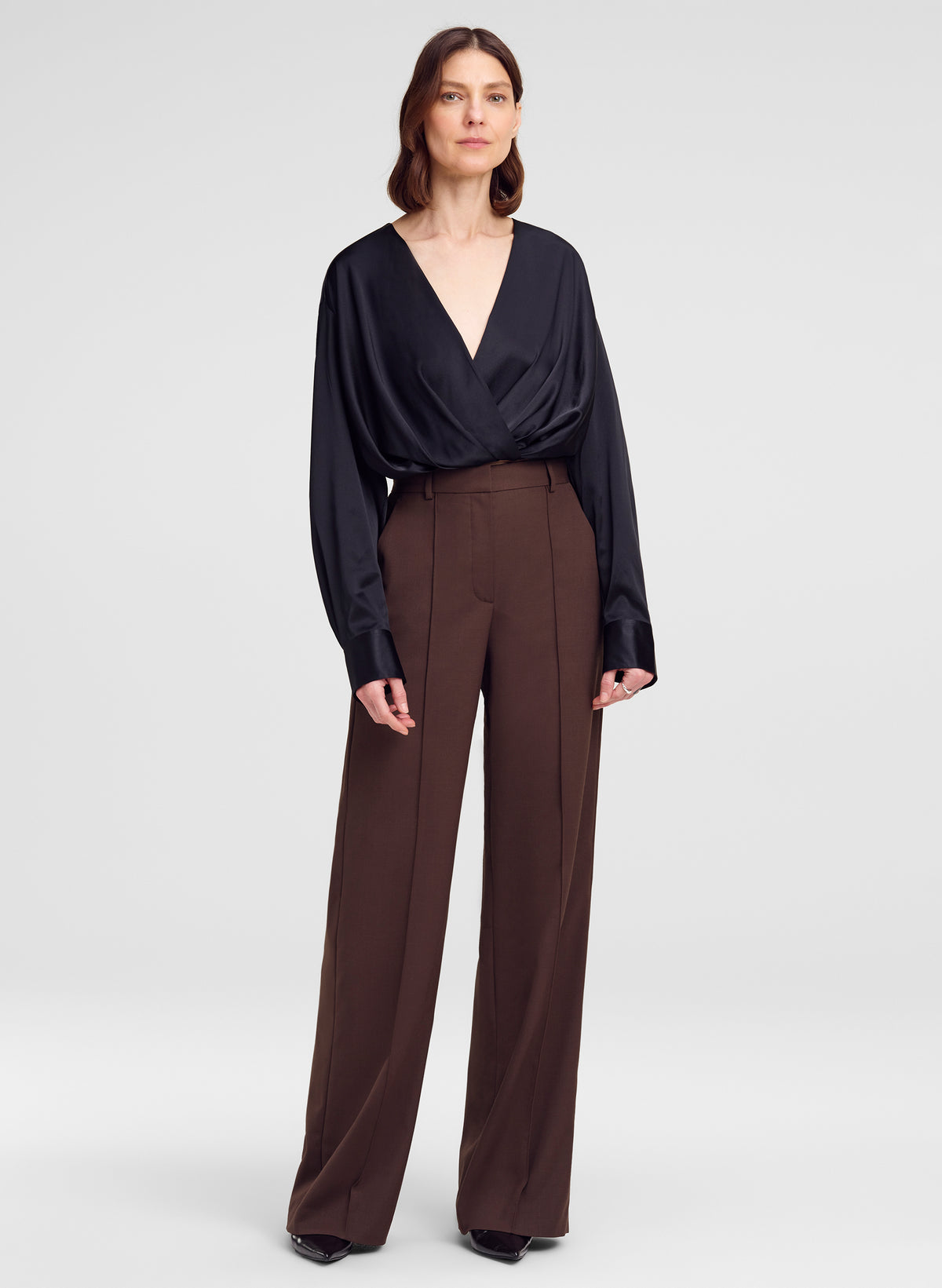 A woman modeling a black long-sleeve wrap-style blouse and high-waisted brown wide-leg trousers against a light gray studio background.
