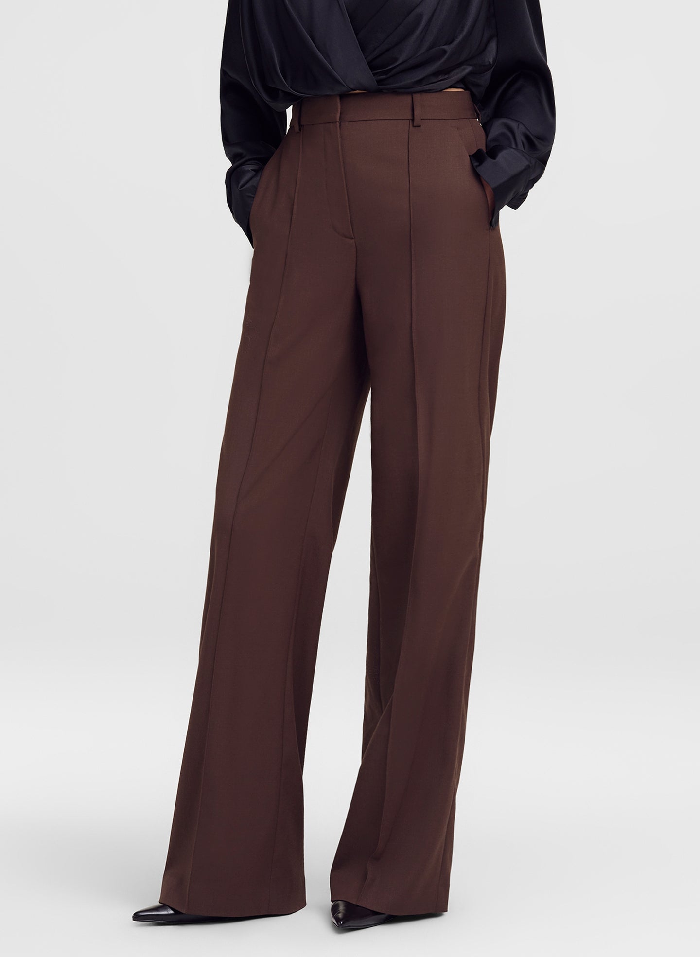 A model wearing high-waisted, wide-leg brown trousers paired with a black satin blouse and black pointed-toe heels, shown from the waist down against a light gray studio background.