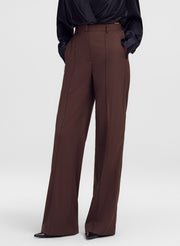 A model wearing high-waisted, wide-leg brown trousers paired with a black satin blouse and black pointed-toe heels, shown from the waist down against a light gray studio background.