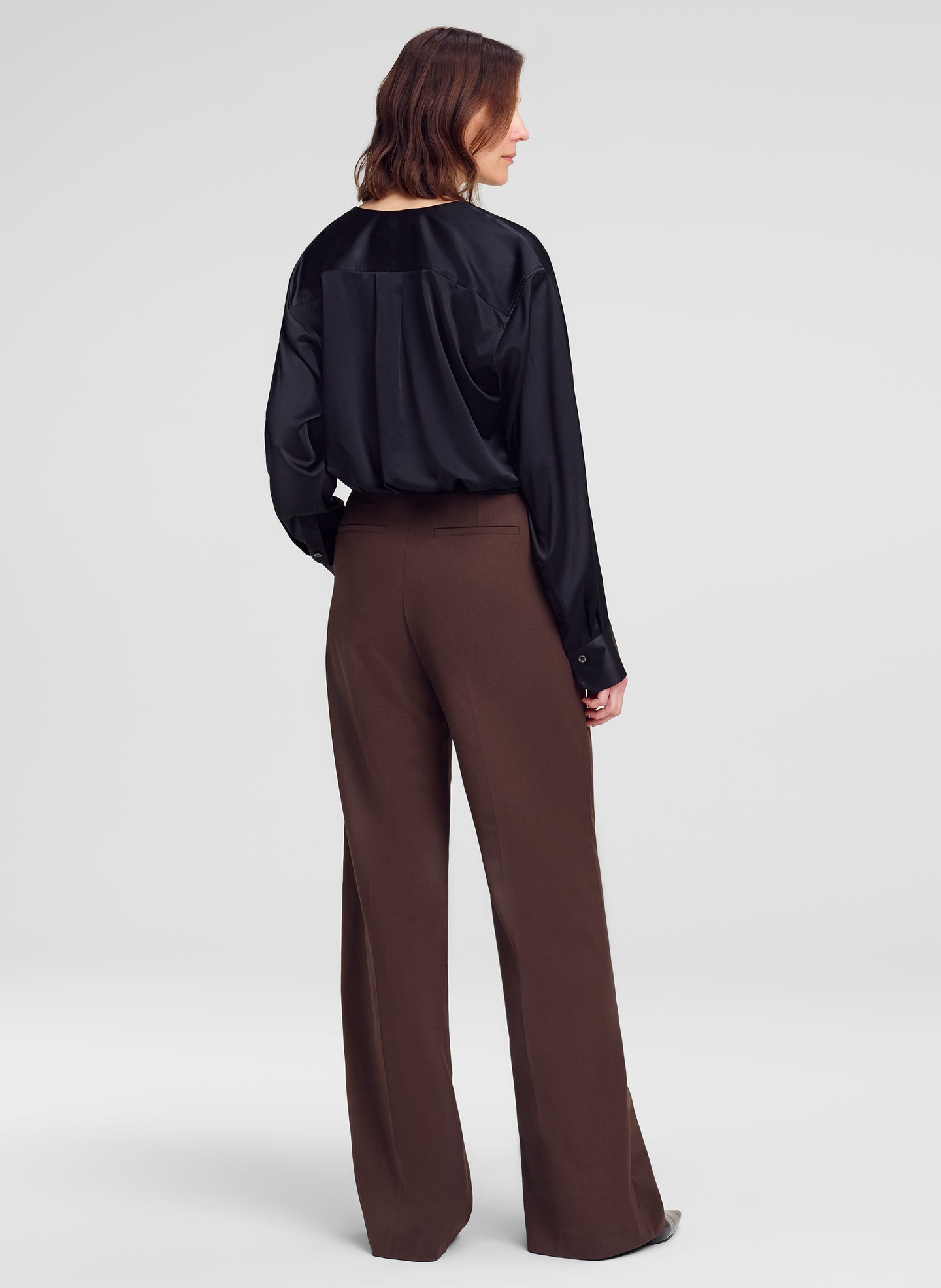 A woman is shown from the back wearing a black satin long-sleeve blouse and brown wide-leg trousers against a light gray studio background.