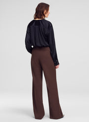 A woman is shown from the back wearing a black satin long-sleeve blouse and brown wide-leg trousers against a light gray studio background.