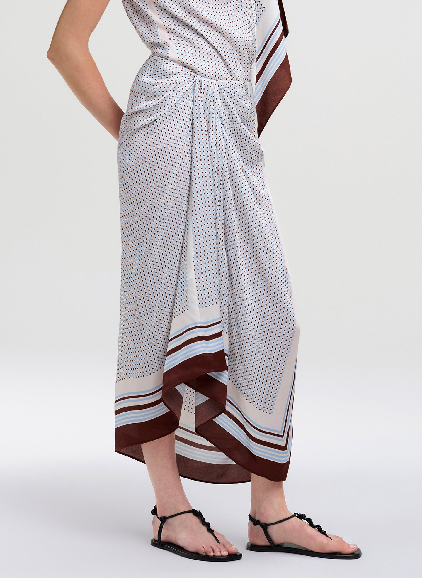A model wearing a white wrap-style midi skirt with a small brown polka dot pattern, brown and white striped borders, and black thong sandals, shown from the waist down against a light gray studio background.