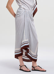 A model wearing a white wrap-style midi skirt with a small brown polka dot pattern, brown and white striped borders, and black thong sandals, shown from the waist down against a light gray studio background.