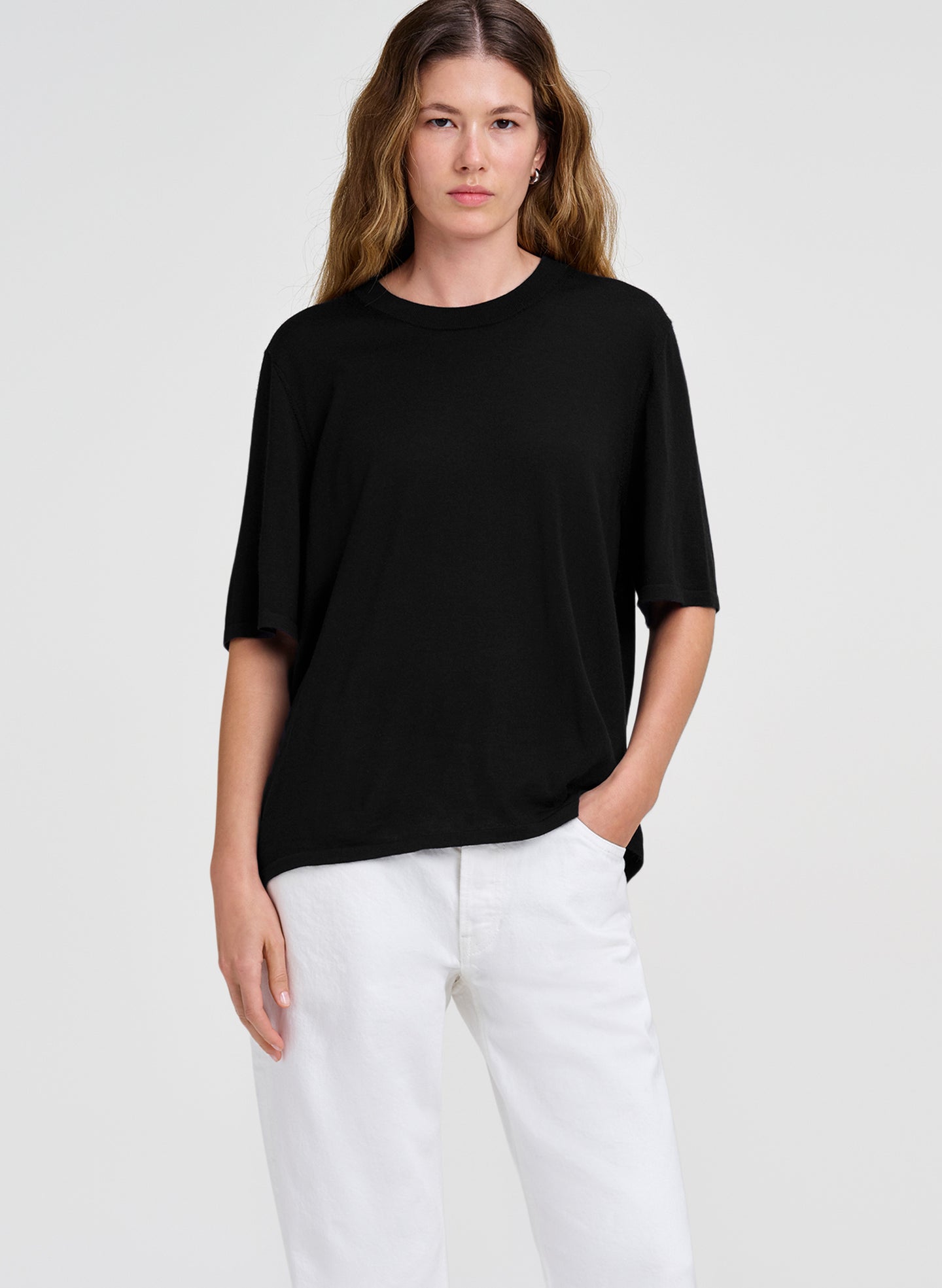 A woman modeling a black short-sleeve crewneck merino shirt paired with white pants, photographed against a solid light gray studio background.