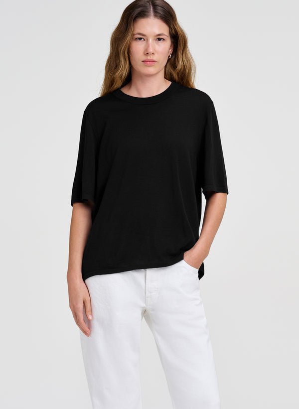 A woman modeling a black short-sleeve crewneck merino shirt paired with white pants, photographed against a solid light gray studio background.