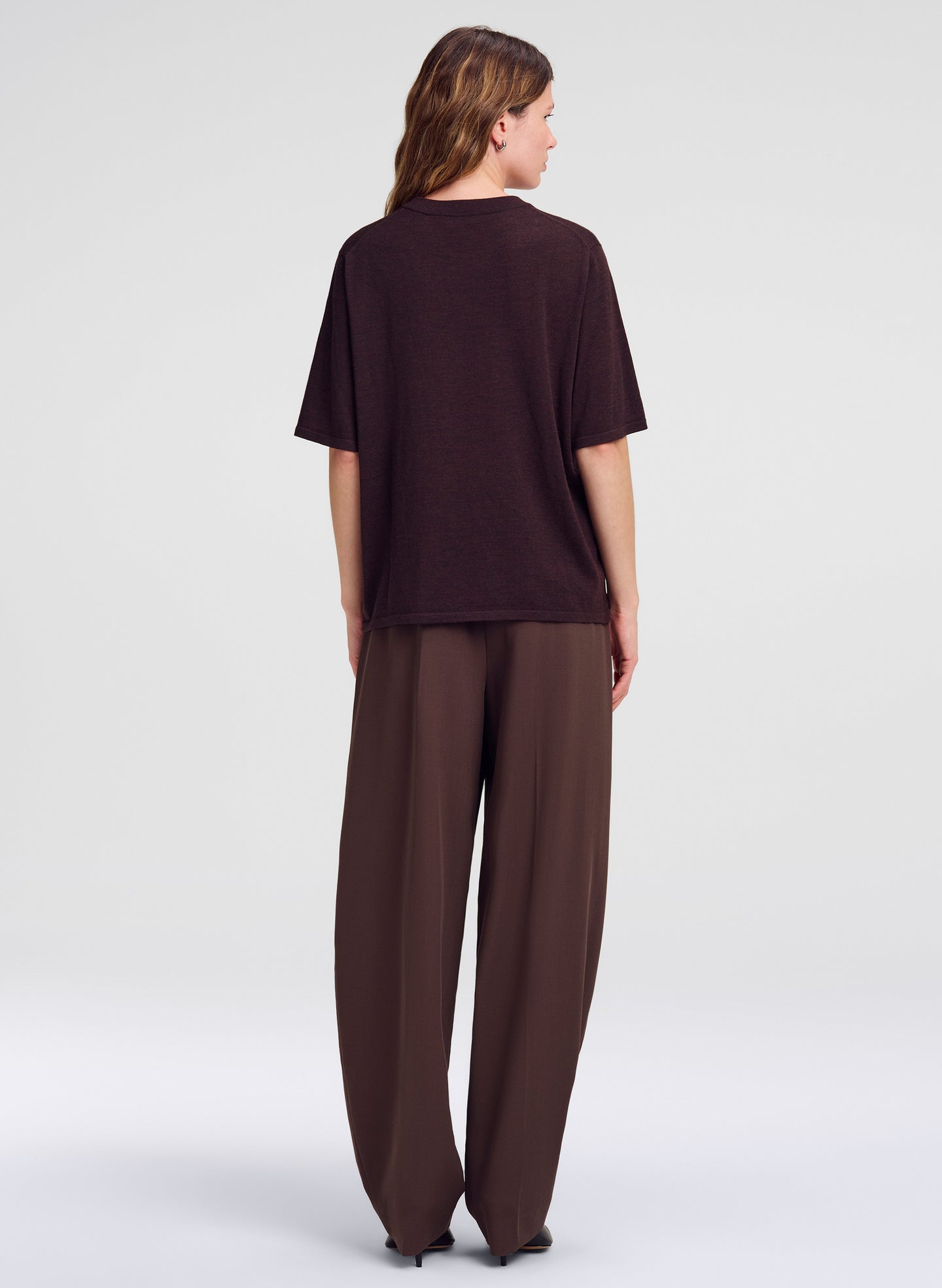 A woman viewed from the back wearing an oversized short-sleeve top and wide-leg trousers in a dark brown color, set against a plain light gray studio background.