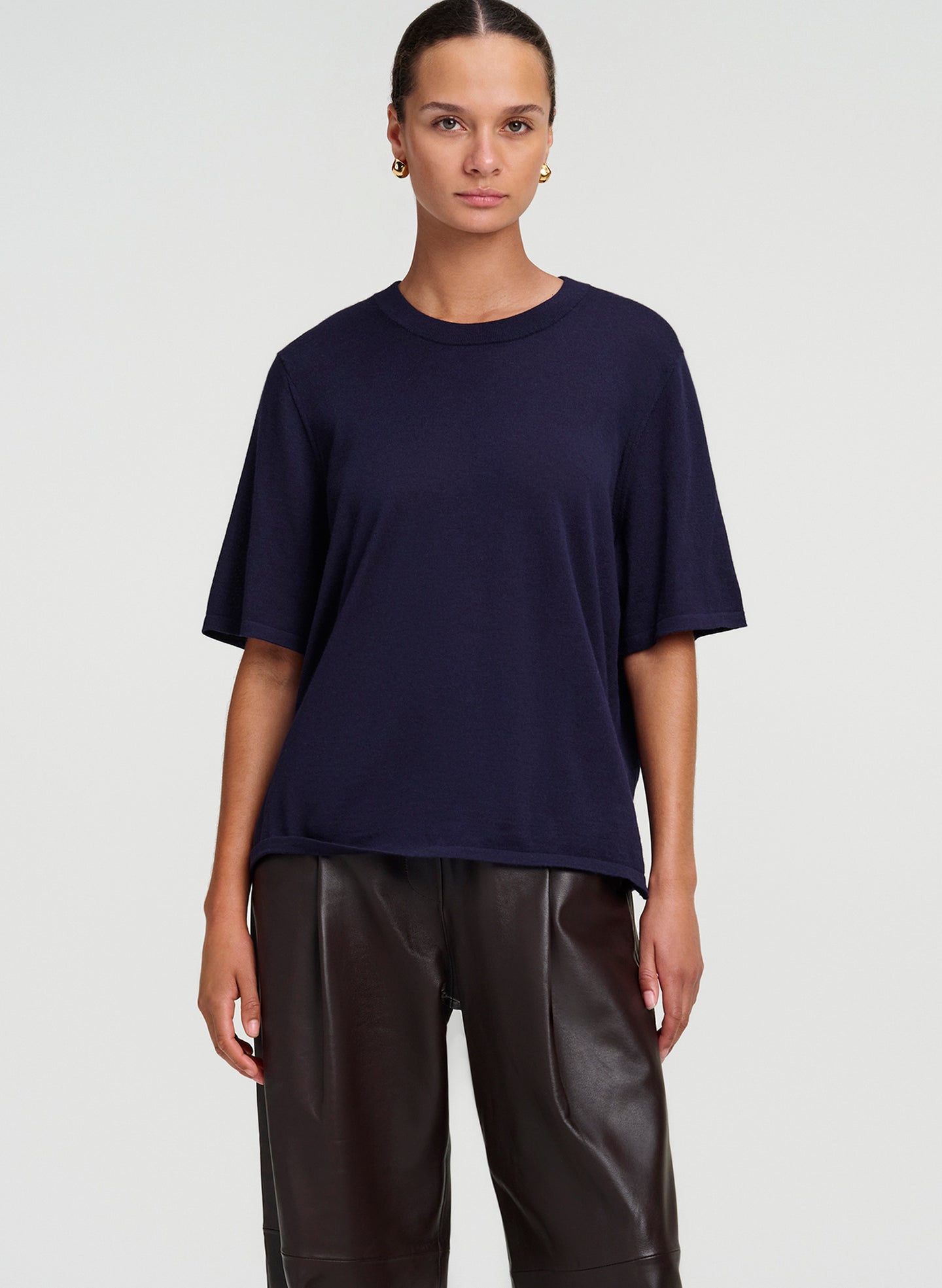 A woman modeling a navy blue short-sleeve crewneck t-shirt paired with dark brown leather pants, shot against a light gray studio background.