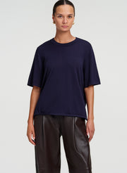 A woman modeling a navy blue short-sleeve crewneck t-shirt paired with dark brown leather pants, shot against a light gray studio background.