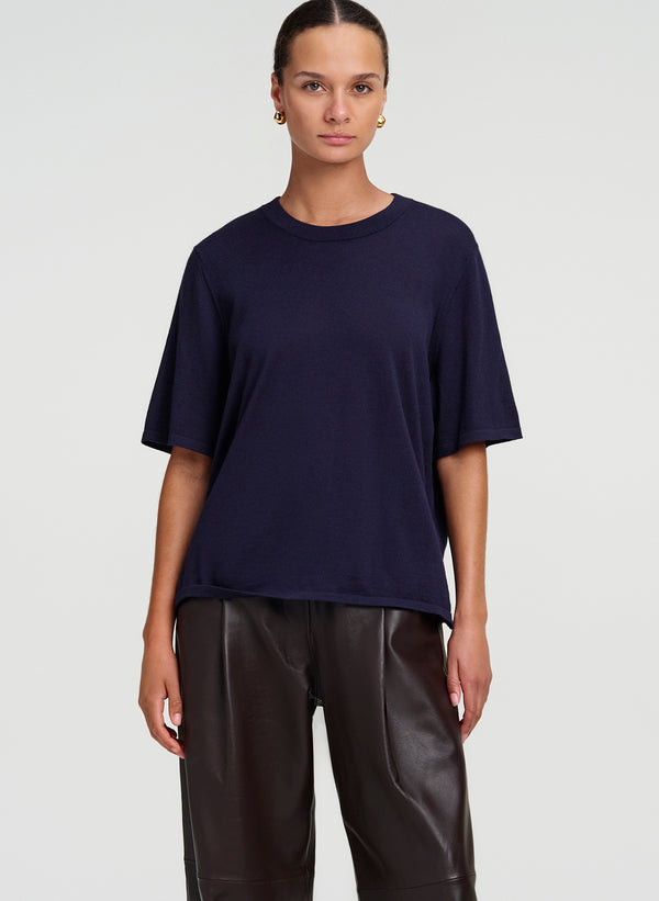 A woman modeling a navy blue short-sleeve crewneck t-shirt paired with dark brown leather pants, shot against a light gray studio background.