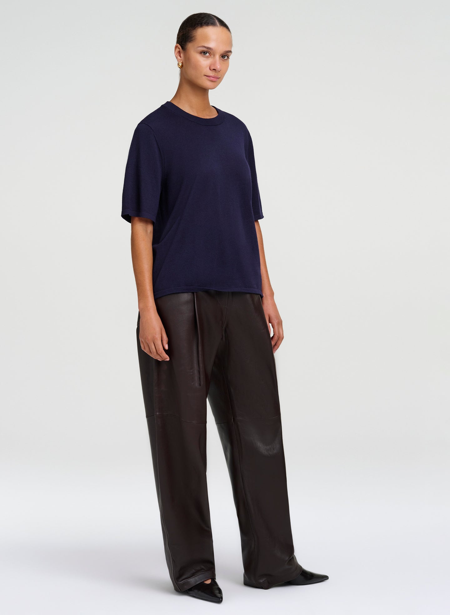 A model wearing a dark navy blue short-sleeve crewneck t-shirt and black wide-leg trousers, standing against a light gray studio background.