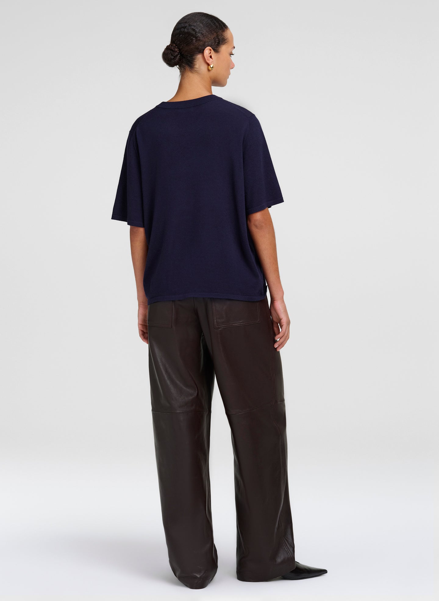 A model shown from the back wearing an oversized navy blue short-sleeve t-shirt and dark brown wide-leg trousers, photographed against a light gray studio background.