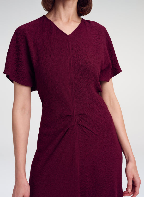 A woman wearing a short-sleeve V-neck dress in deep burgundy with a textured crinkle fabric and a gathered twist detail at the waist, shown against a light gray studio background.