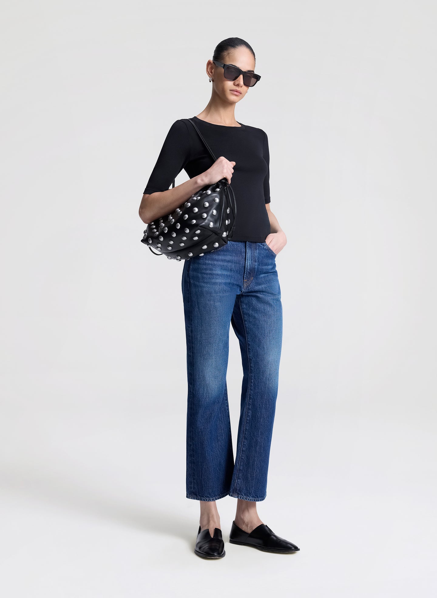 Woman wearing a black top and blue jeans, holding a studded handbag on a white background