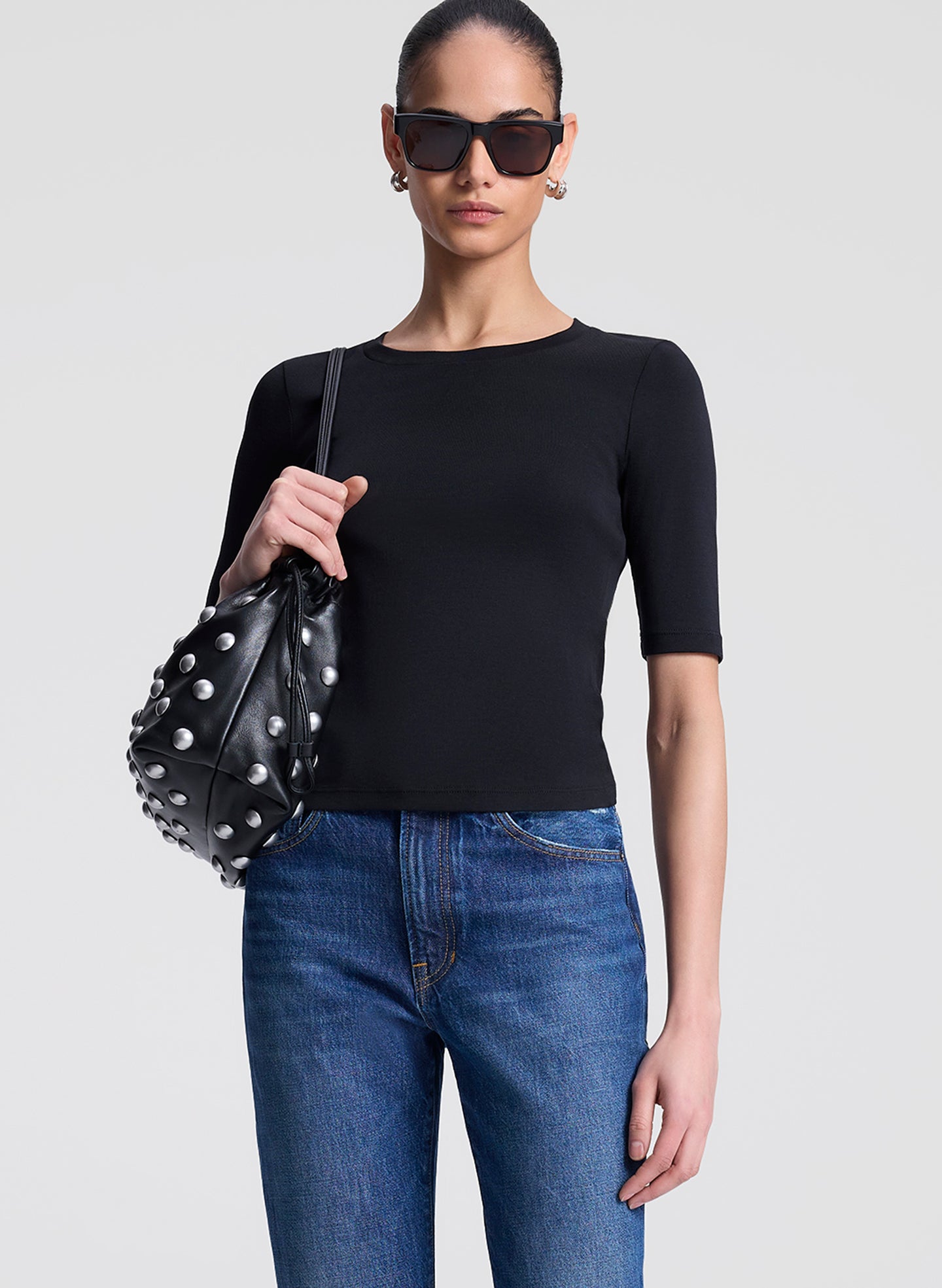 Woman wearing a black top and blue jeans holding a black studded handbag against a light gray background