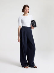 Woman wearing a white top and navy pants holding a black clutch against a white background