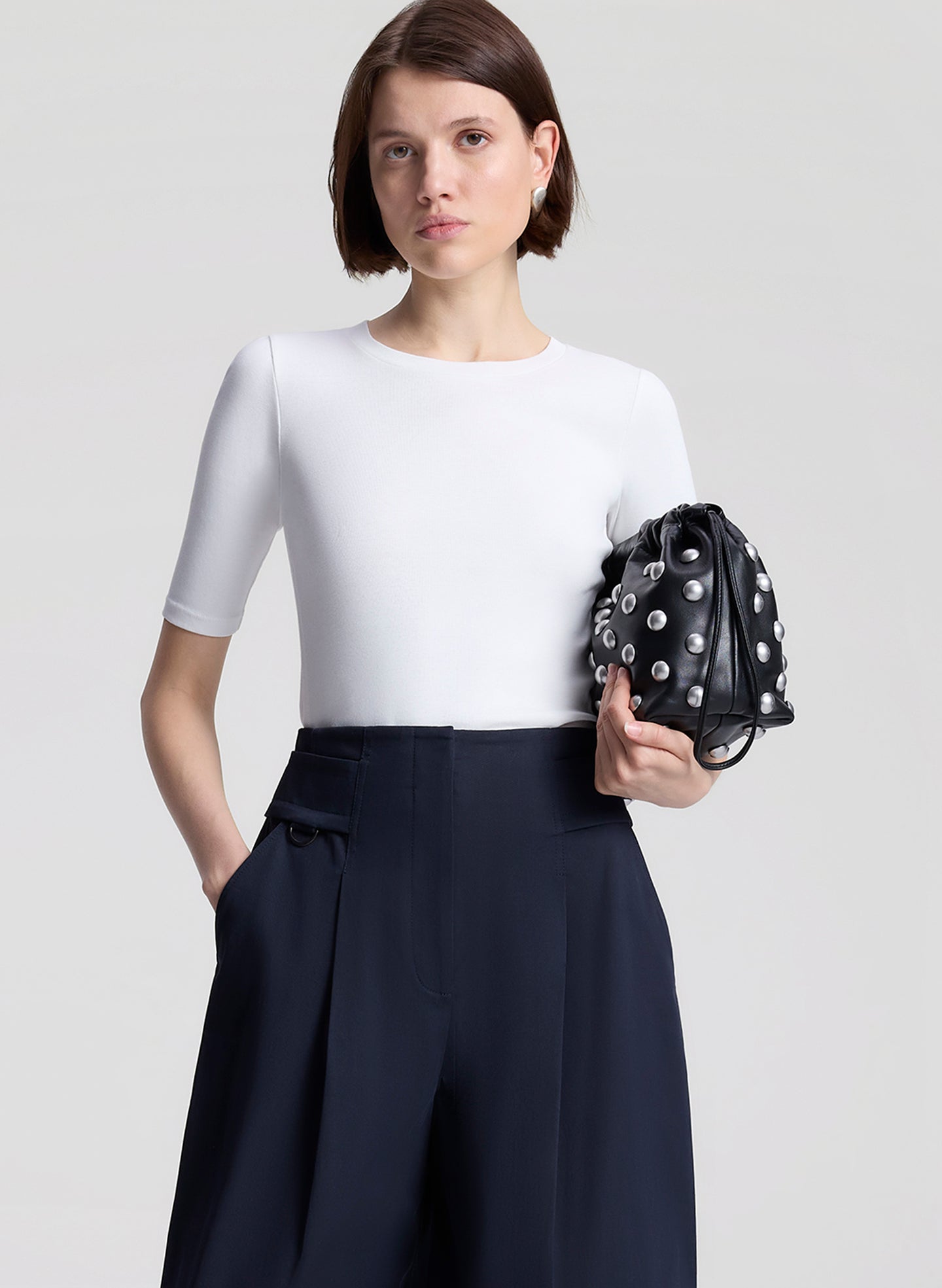 Woman wearing a white top and navy pants holding a black studded handbag on a light gray background