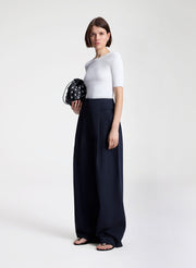 Woman wearing a white top and navy wide-leg pants holding a black clutch against a white background