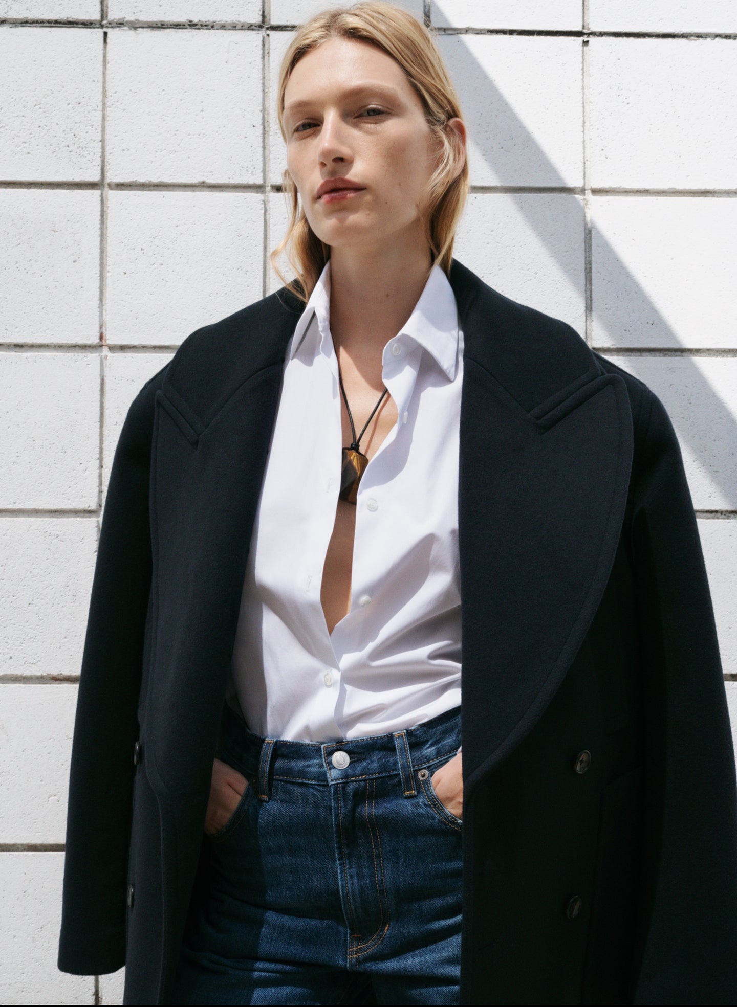 Person wearing a navy blue coat over a white shirt and blue jeans against a light-colored wall.