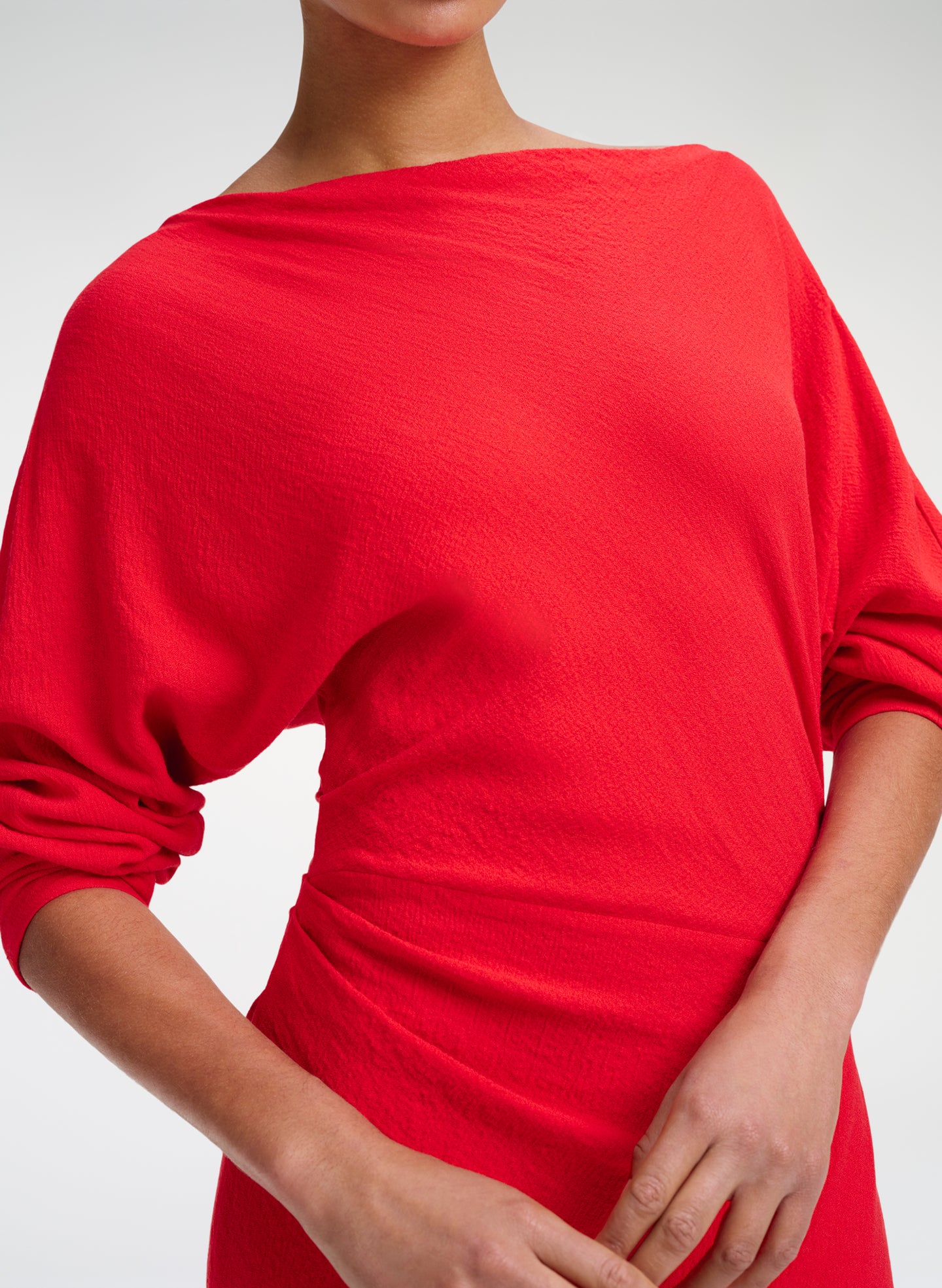 A close-up of a model wearing a bright red, boat-neck knit top with draped, elbow-length sleeves and a gathered waist detail, set against a plain light gray background.