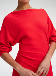 A close-up of a model wearing a bright red, boat-neck knit top with draped, elbow-length sleeves and a gathered waist detail, set against a plain light gray background.